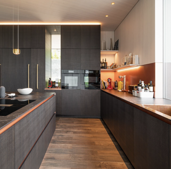 Küche der Käslin Innenausbau AG in Steinhausen mit dunklen Schrankfronten, Holzarbeitsplatte und warm beleuchteter Rückwand.